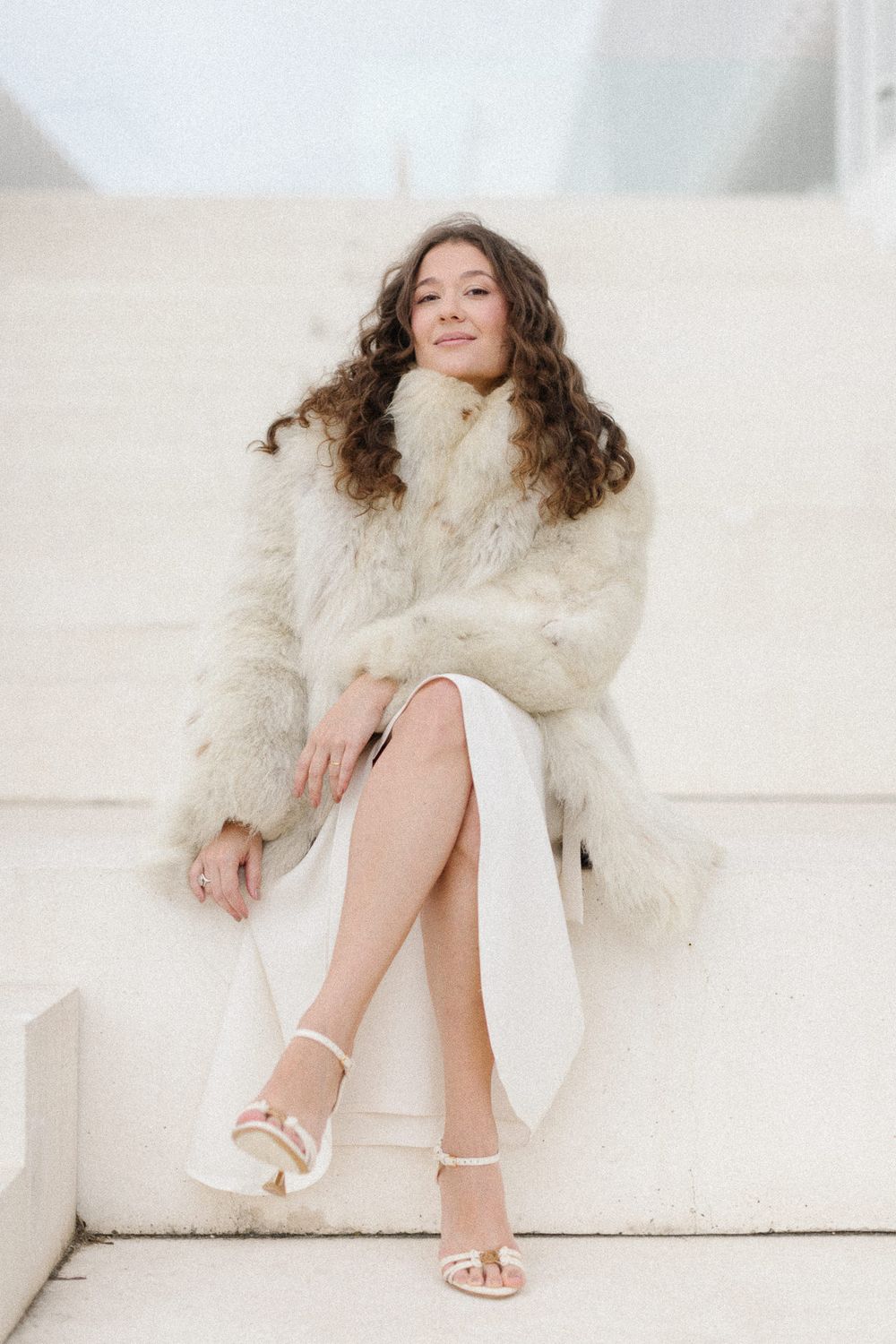 A person in a fluffy white coat lounges elegantly on a white couch wearing strappy sandals.