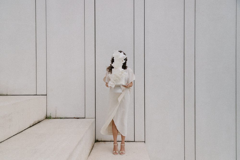 A figure in a white dress stands against a minimalist grey wall with geometric lines.