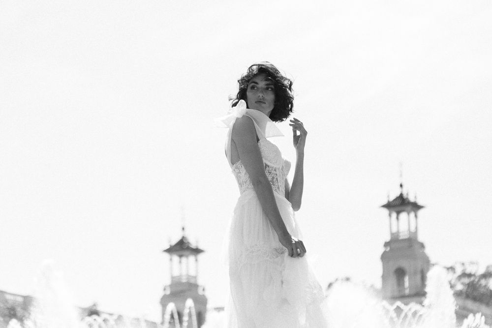 Black and white photo of an elegant woman in a white dress posing gracefully outdoors near ornate architecture.