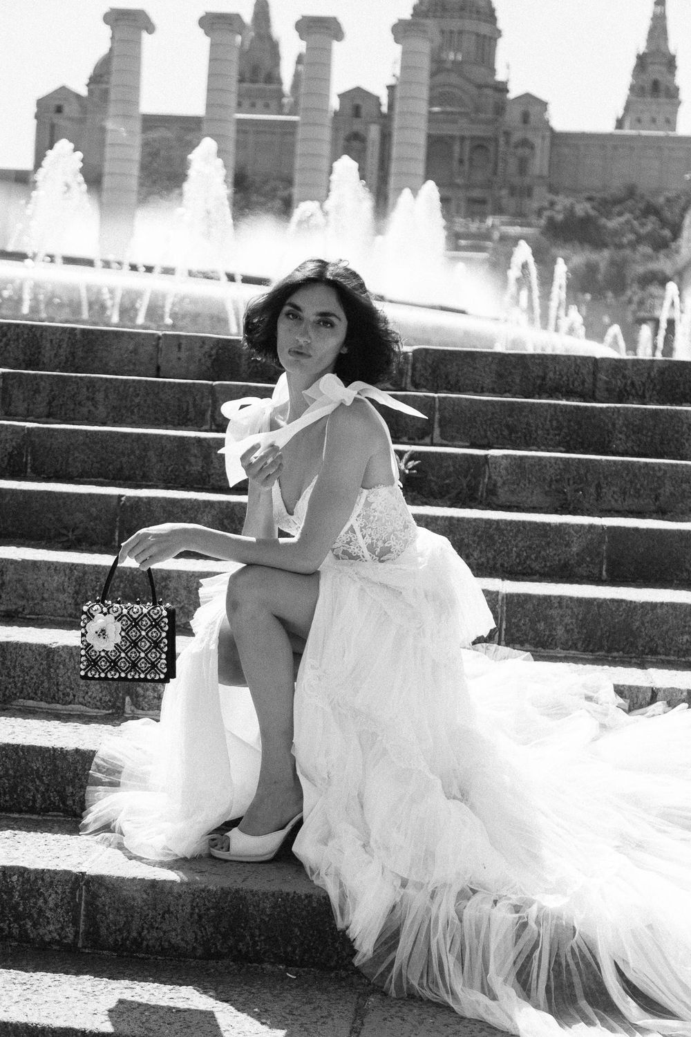 Black and white photo of a bride in a ruffled white gown sitting on steps near a fountain.