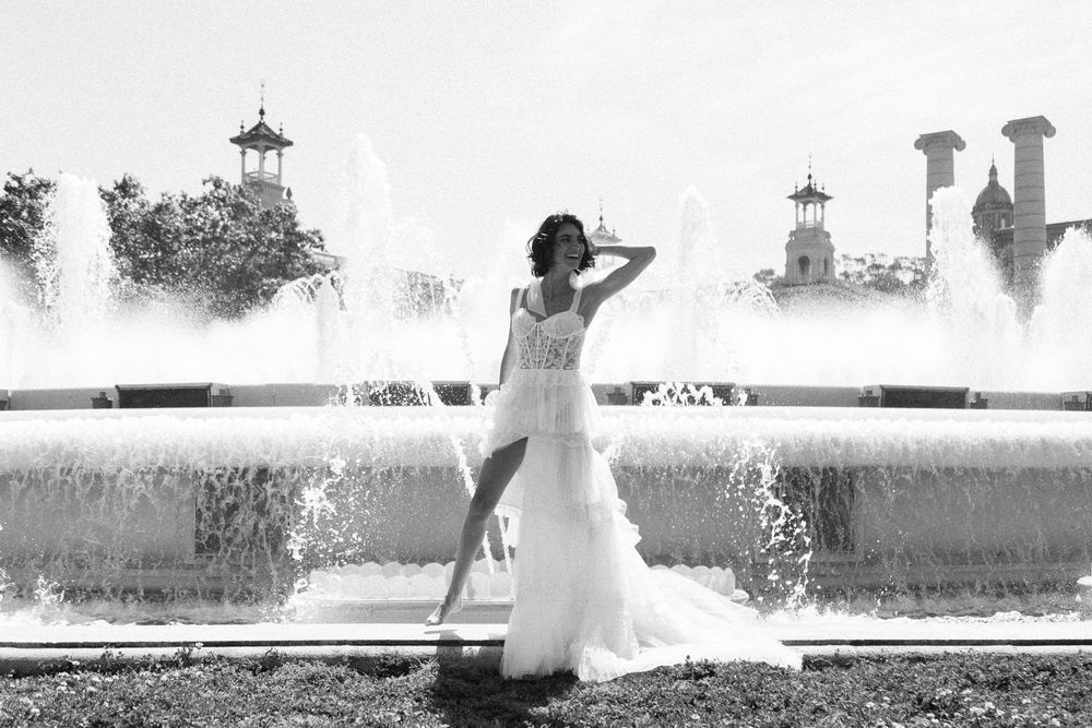 Woman in elegant white wedding gown poses gracefully before grand fountain in black and white photo.