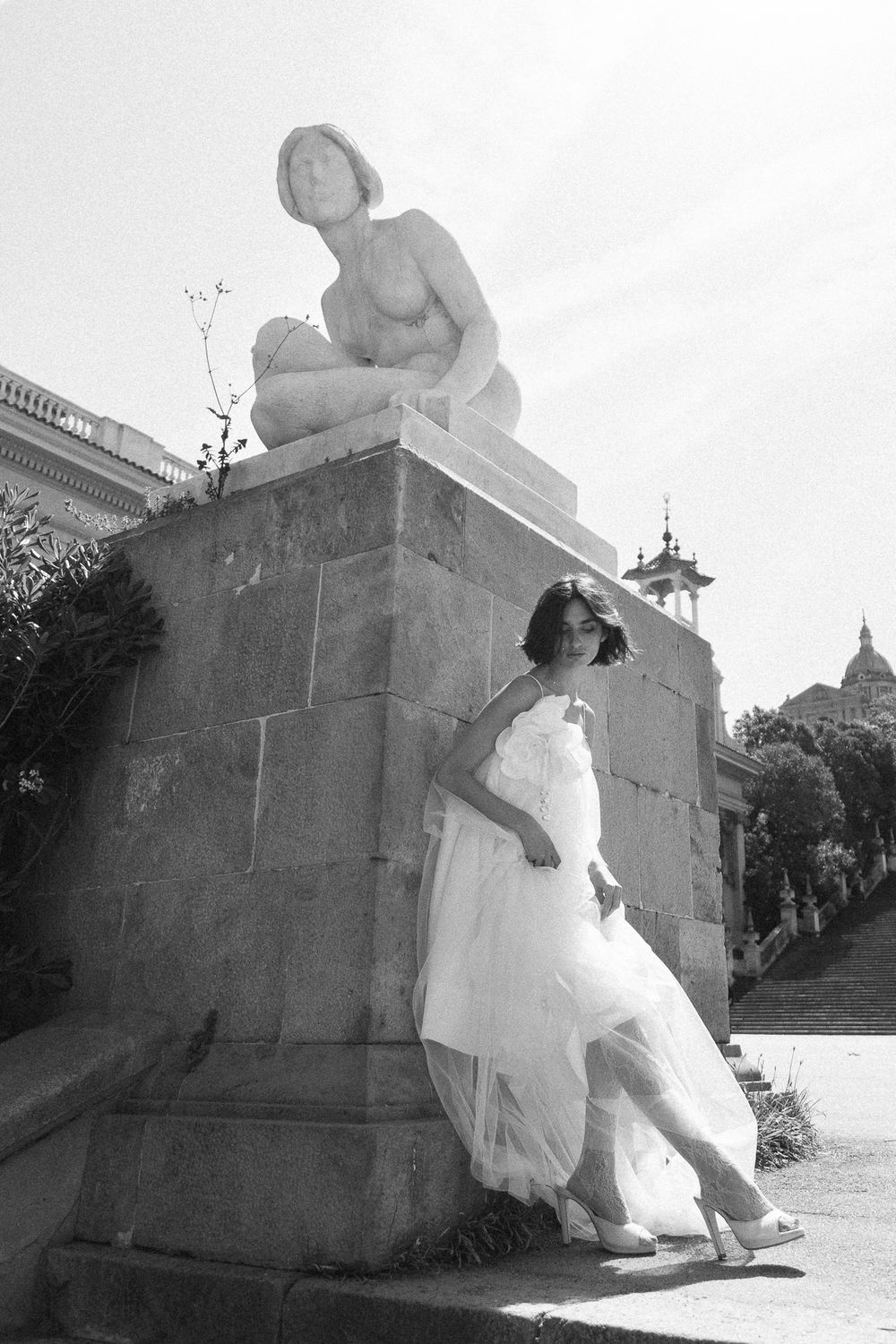 Black and white photo of a bride in white dress leaning against a stone statue pedestal outdoors.