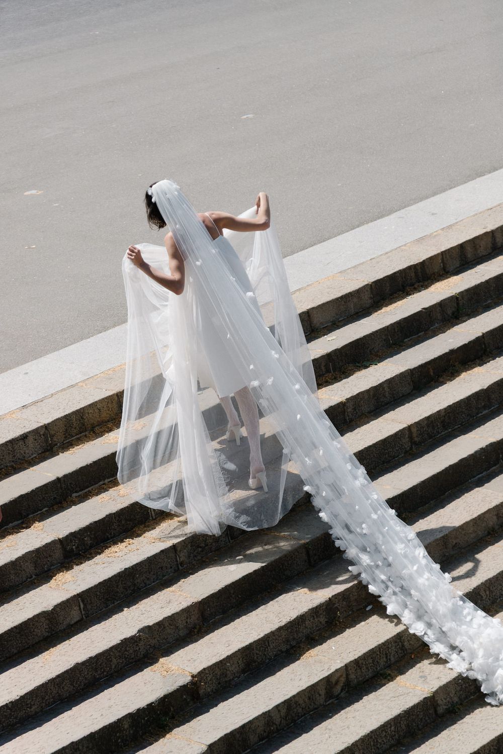 Bride in white wedding dress and long floral veil descending outdoor stone steps on a sunny day.