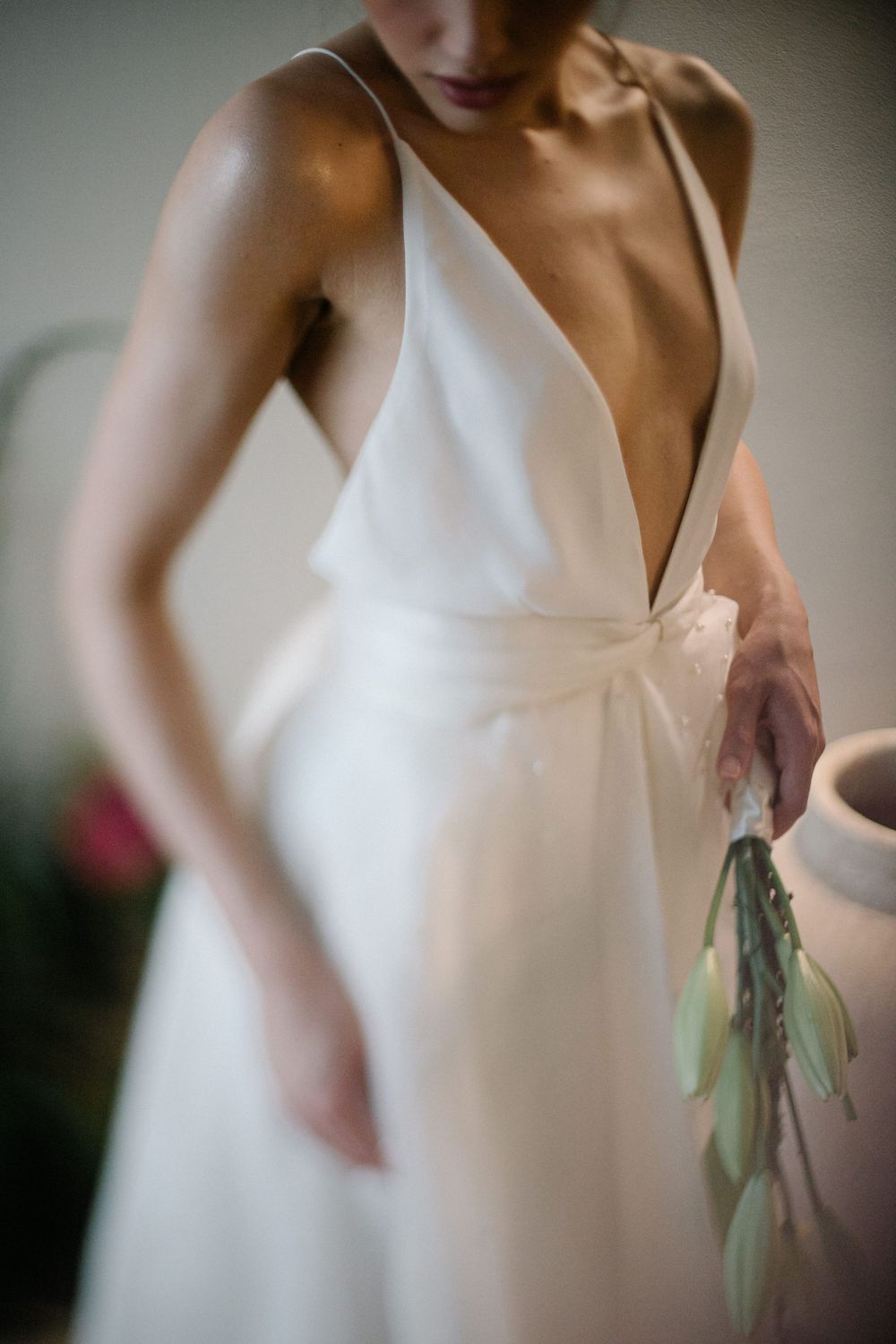 Bride in elegant deep V-neck white wedding gown holding greenery bouquet, soft focus bridal portrait.