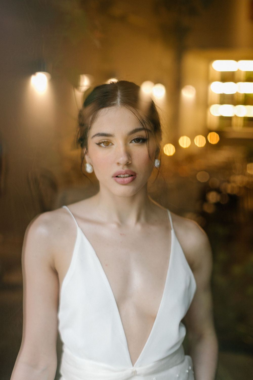 Woman in white deep-V wedding gown poses elegantly with bokeh lights in background.