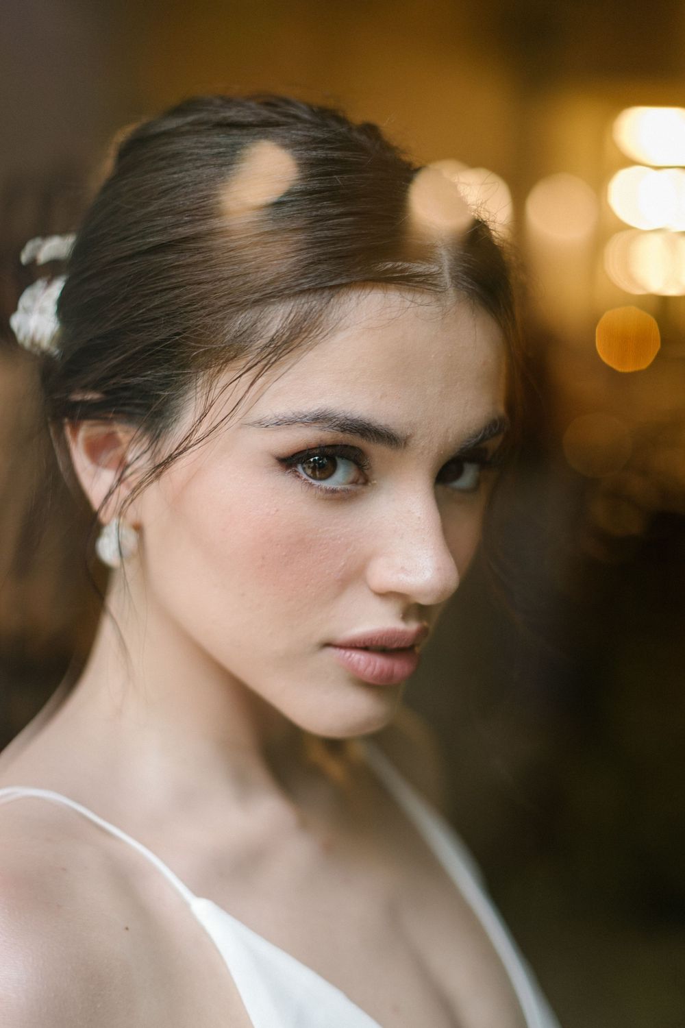 Young brunette bride with elegant updo, white flower hair accessory, pearl earring, and natural makeup gazes softly at camera.