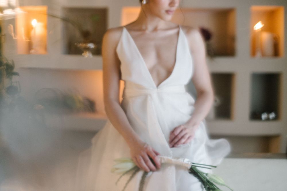 Bride in elegant deep V-neck white wedding gown holding a green bouquet in a softly lit room.