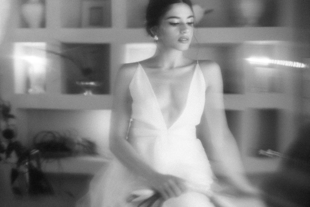 Black and white photo of an elegant woman in a white V-neck dress, seated gracefully in a softly lit room.