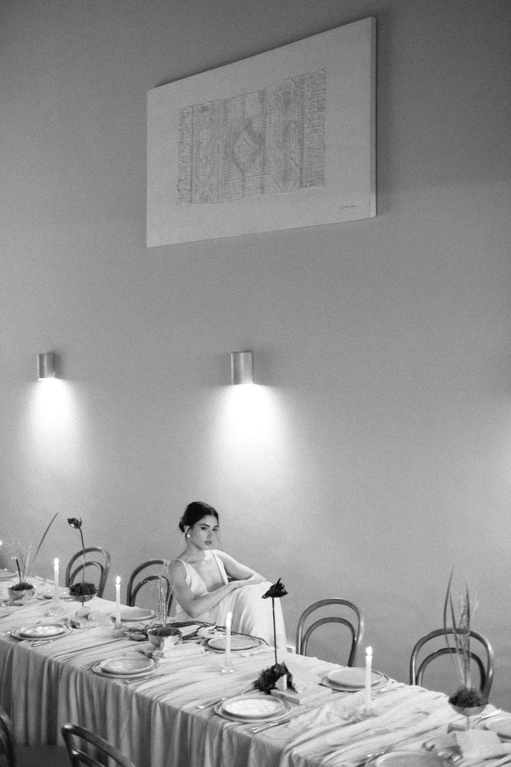 Black and white photo of a woman seated alone at an elegantly set dining table with candles and wall sconces.