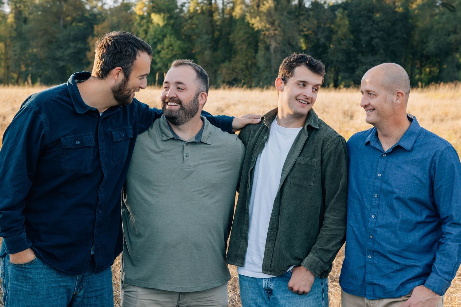 Four adult men laughing and posing together in a sunlit golden field.
