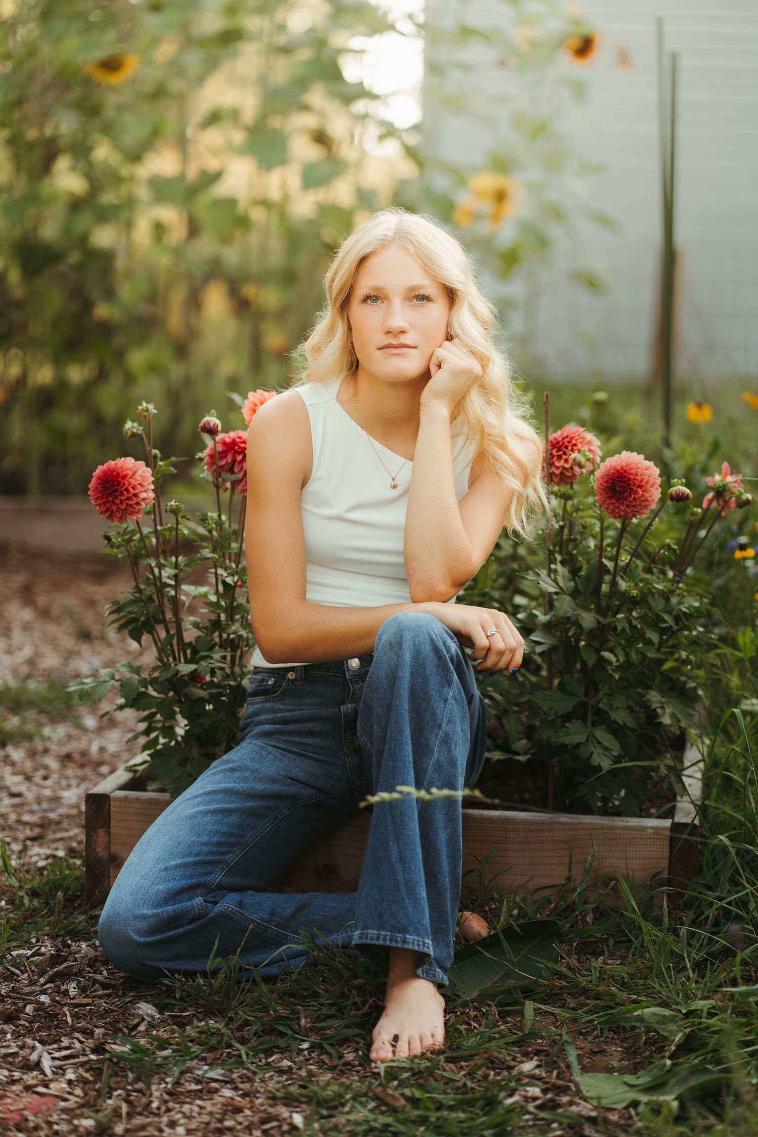 Blonde teen girl in white tank and jeans sitting barefoot in garden surrounded by dahlia flowers.