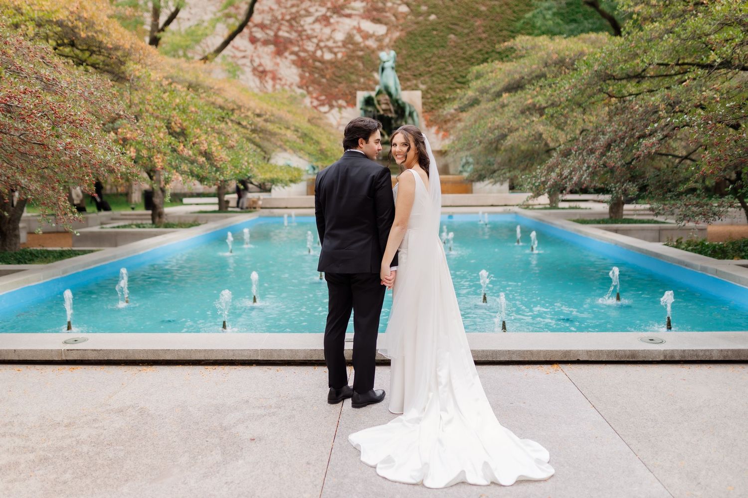 A couple in wedding attire stands near a serene turquoise pool surrounded by autumn trees and foliage.