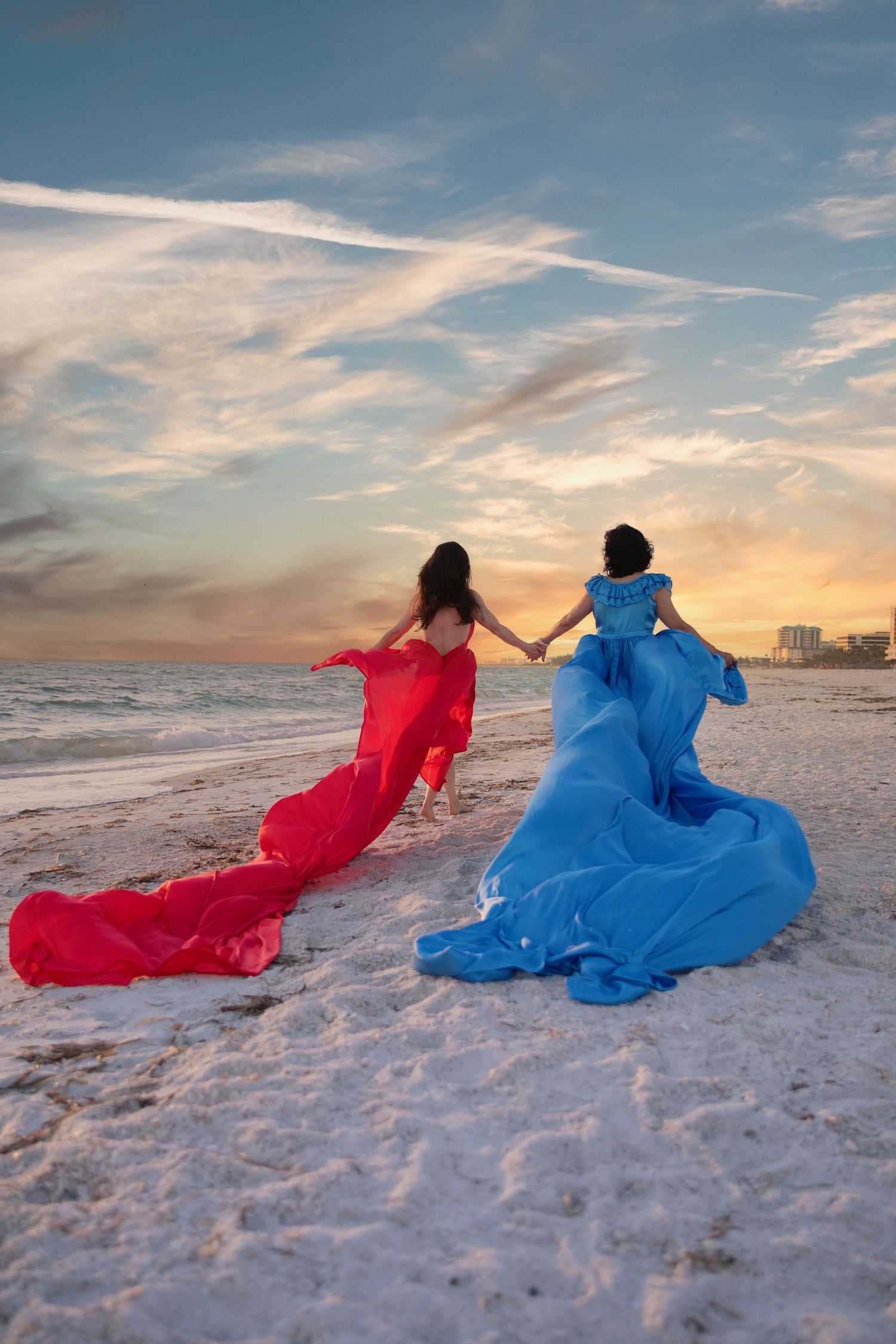 A flowing red ballgown dress billows dramatically against a white building exterior at sunset with palm trees in view.