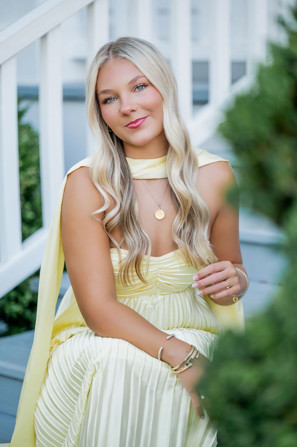 Woman in light yellow dress poses on porch with white railing and green foliage.