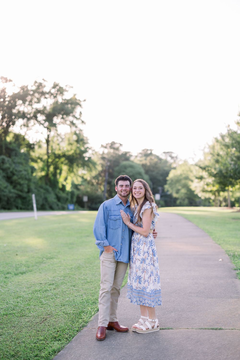 A couple shares a romantic moment together on a winding path surrounded by green grass and trees at sunset.