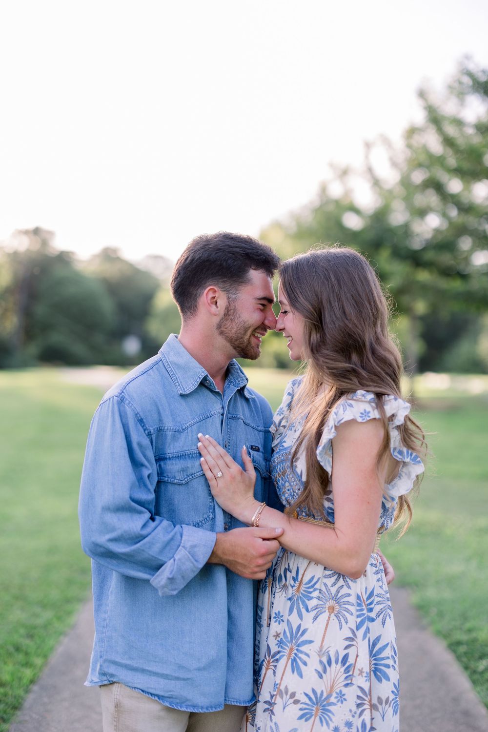 A romantic couple embraces while standing on a path surrounded by green fields at sunset.