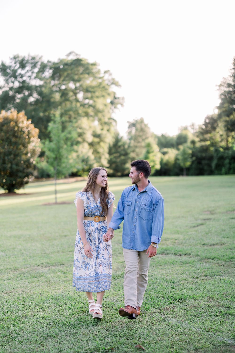 A couple walks together on a grassy hillside during golden hour, wearing casual blue and floral outfits.