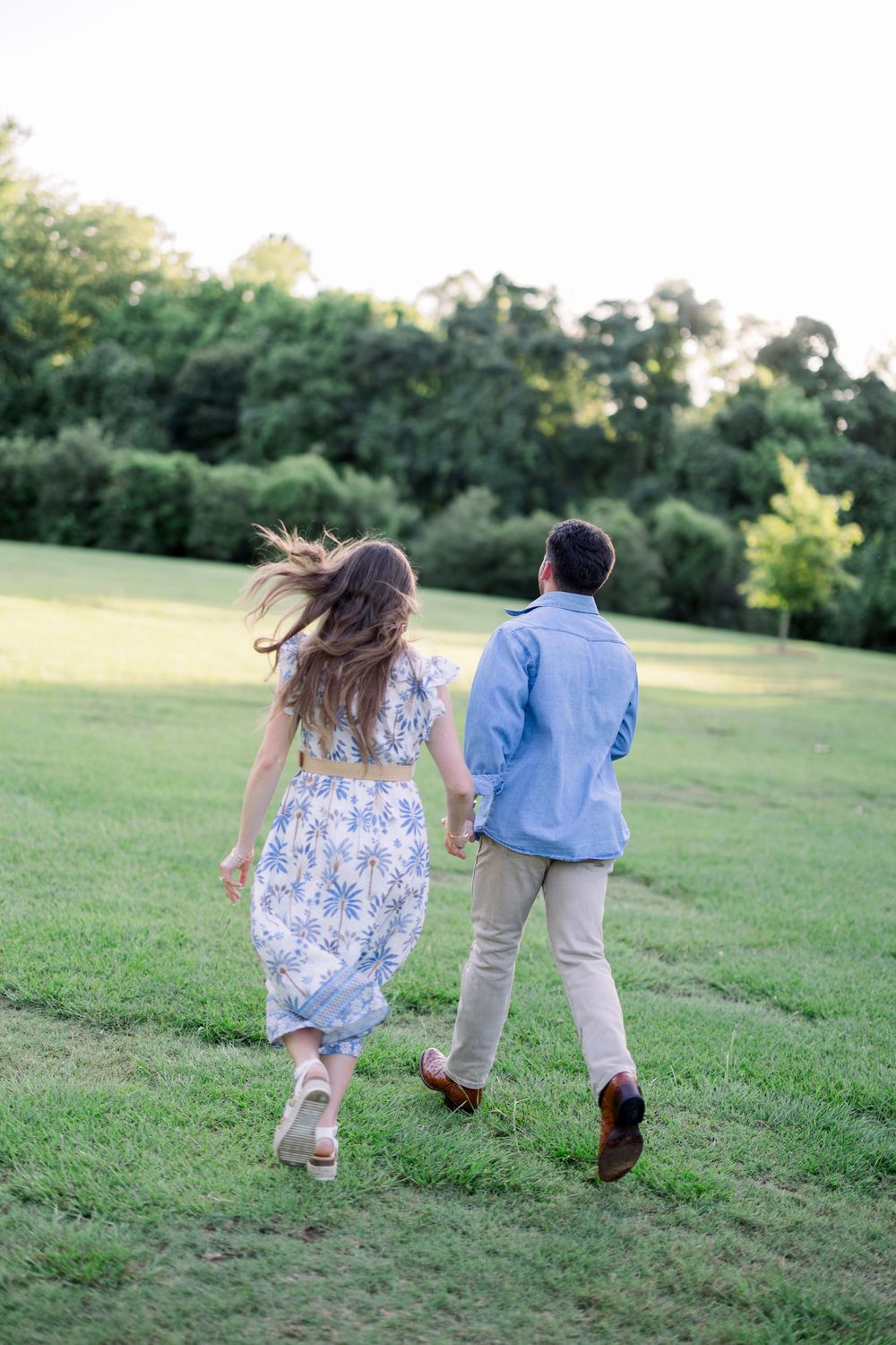 A couple runs playfully up a grassy hill together at sunset, her floral dress flowing in the breeze.
