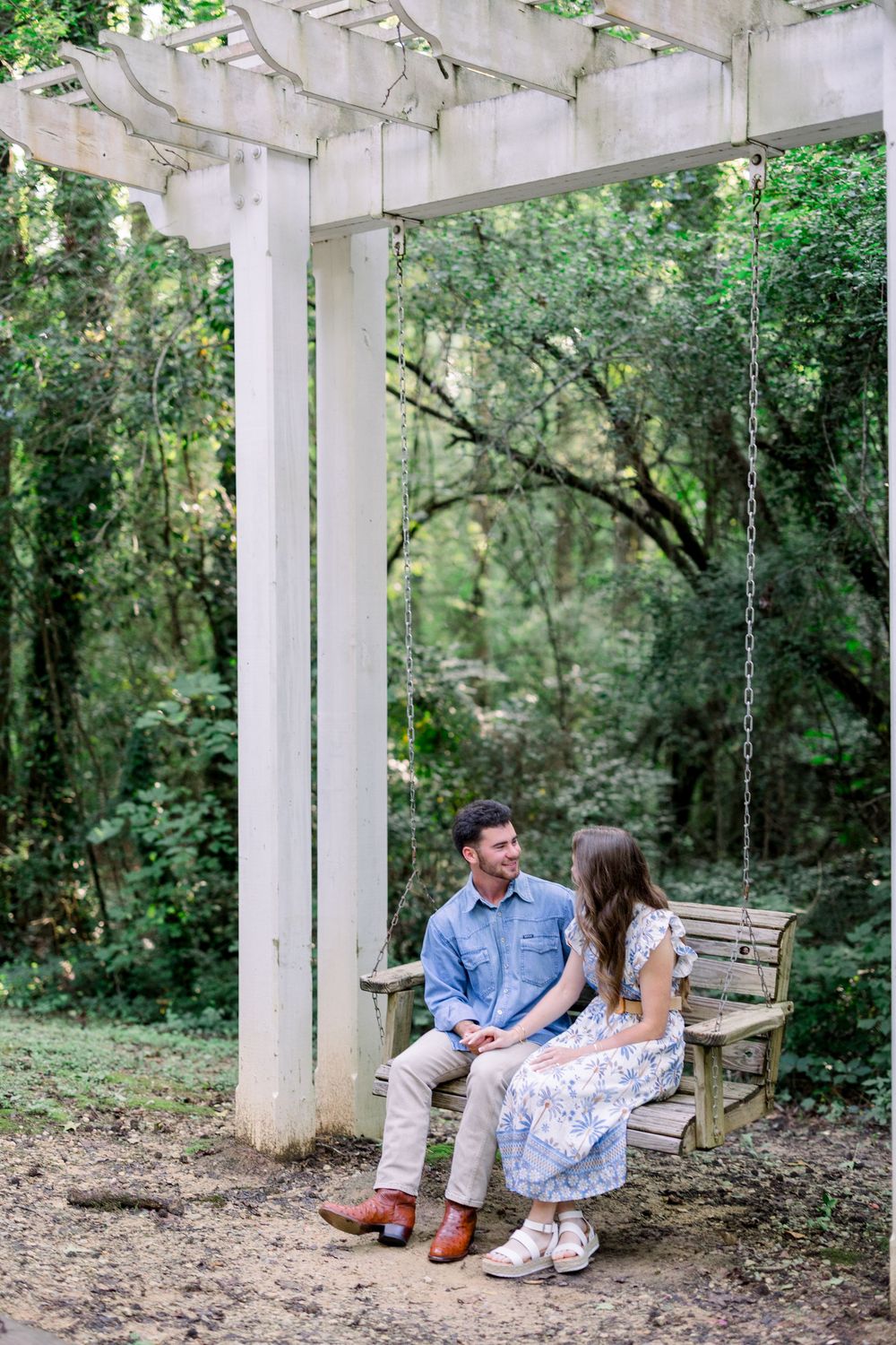 A couple sits together on a rustic white bench surrounded by lush greenery in a garden setting.