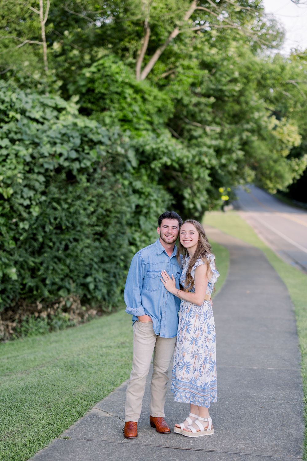 A couple stands together on a tree-lined path during a summer engagement photoshoot.