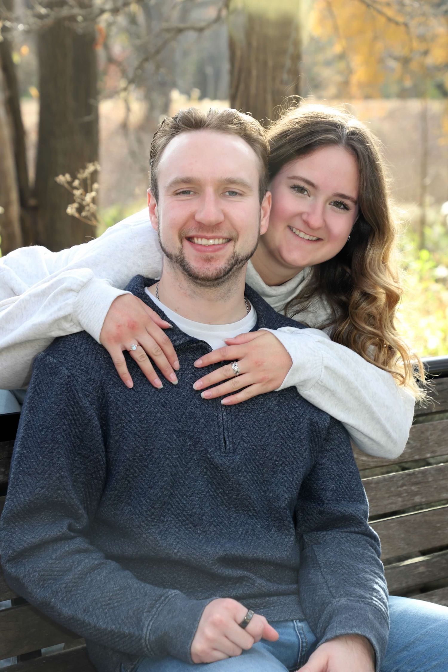 Happy couple posing outdoors on a bench, woman hugging man from behind, both smiling, wearing casual sweaters in a park.