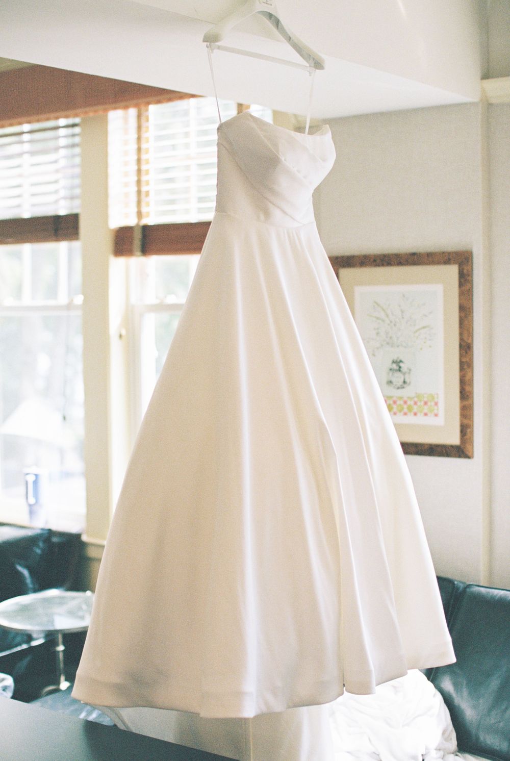 Elegant white satin wedding gown with sweetheart neckline and A-line silhouette hanging in sunlit room with teal furniture.