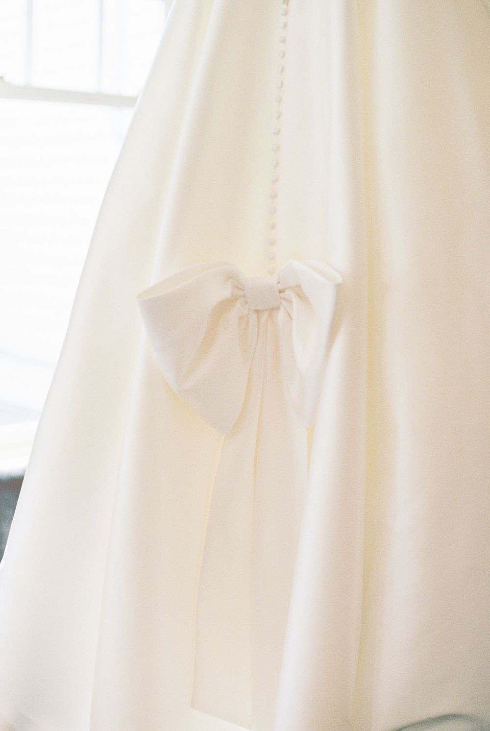 A delicate white bow detail adorns a flowing white wedding dress fabric near a window.