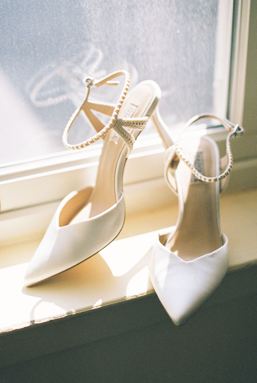 Elegant white bridal heels with pearl ankle straps sit on a windowsill in soft sunlight.