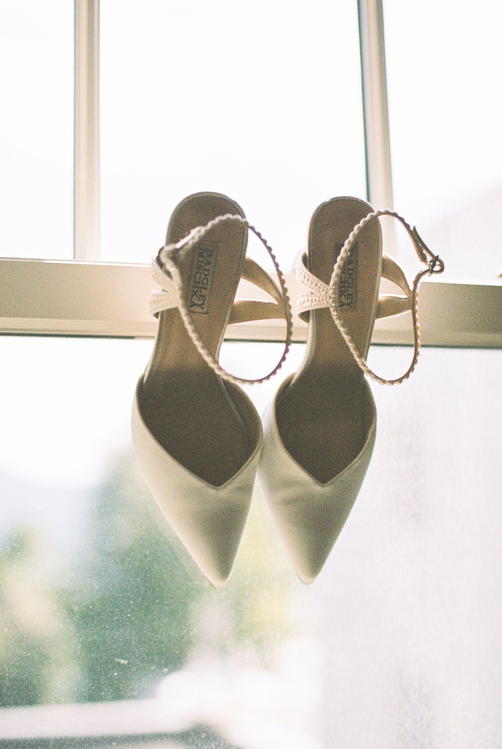 Elegant white pointed-toe heels with delicate ankle straps photographed against a bright window.