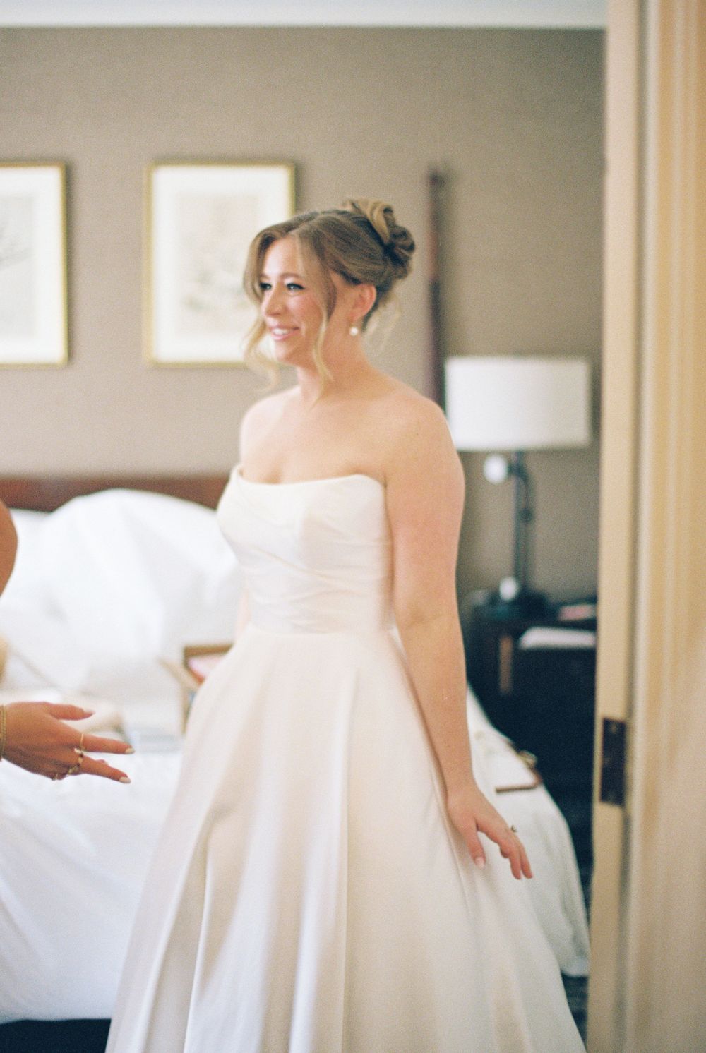 A bride in a flowing white wedding dress being helped with final preparations in an elegant indoor setting.