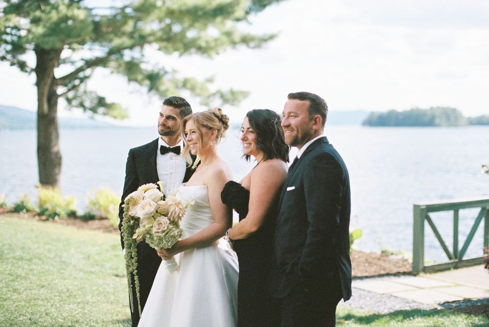 A scenic outdoor wedding ceremony takes place on a grassy lawn overlooking a serene lake or waterfront.
