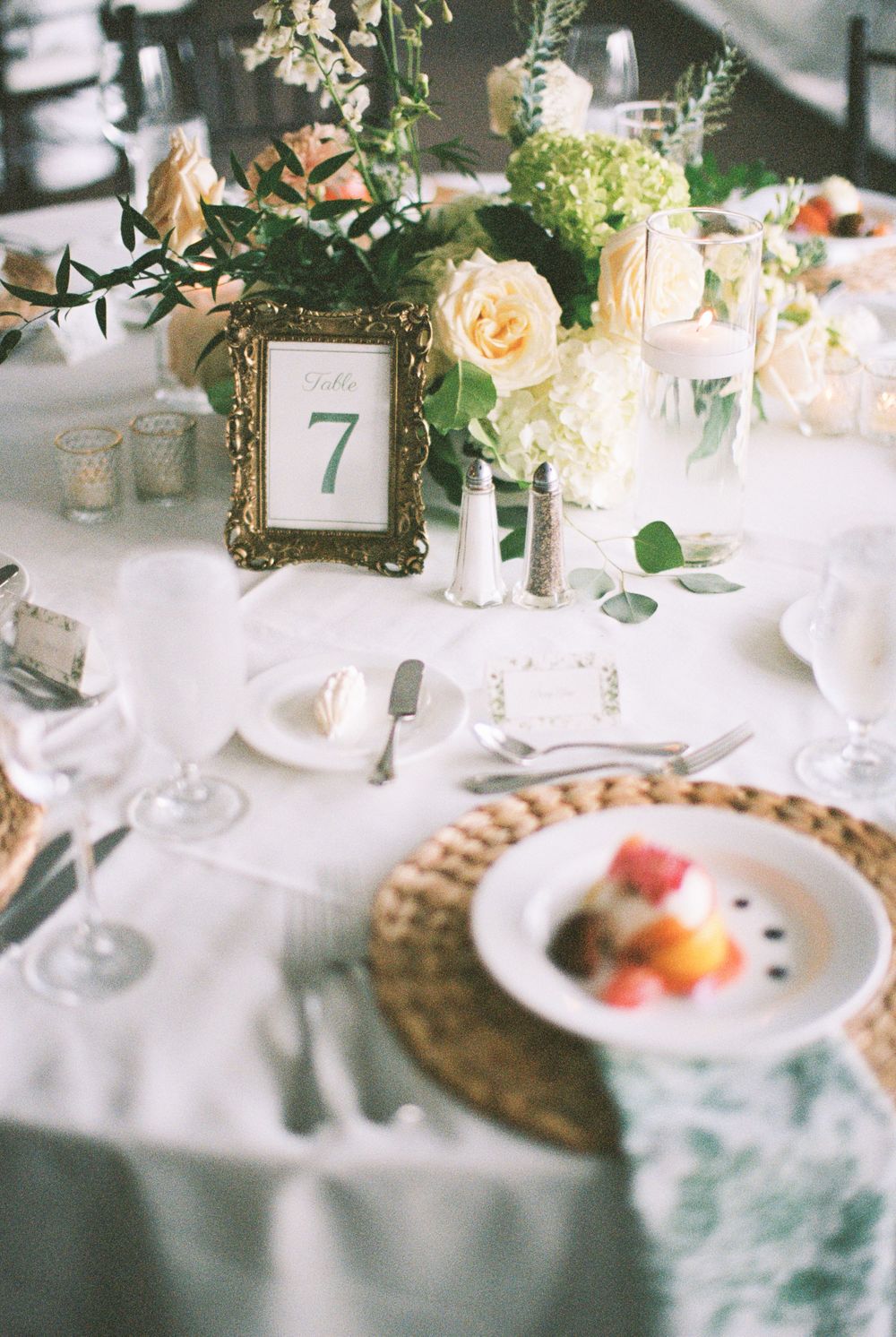 Elegant wedding table setting with gold-framed table number, peach roses, and crystal glassware on white linens.