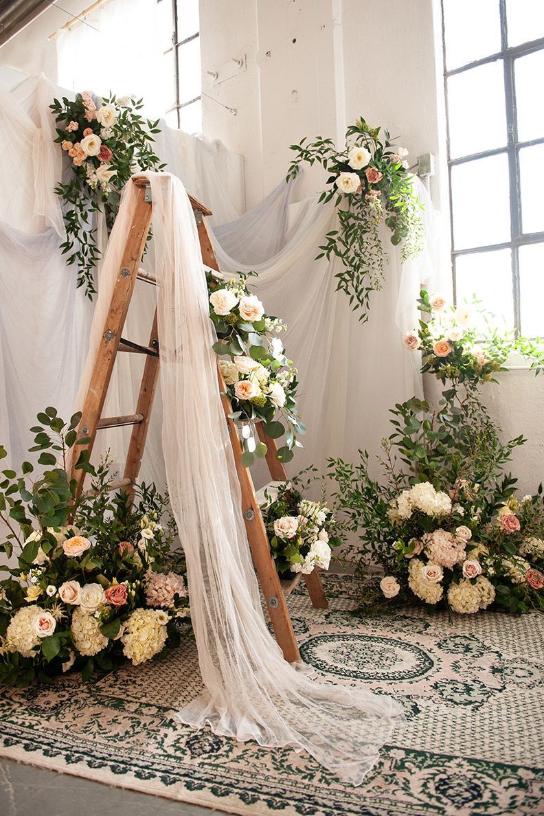 Rustic wooden ladder decorated with flowing white fabric, roses and greenery against a patterned rug in a bright studio space.