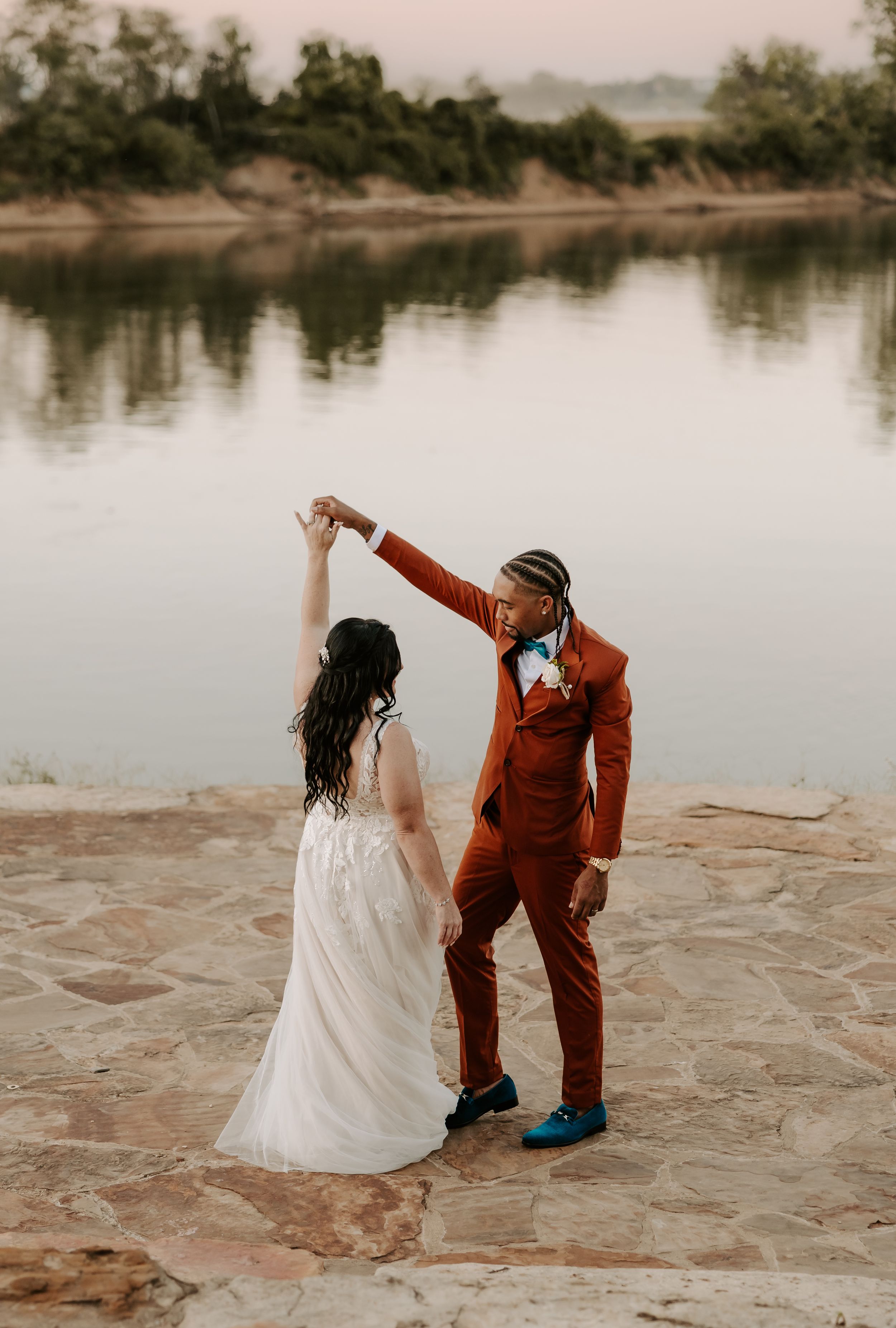 Couple dancing on stone patio by serene lake during sunset wedding photoshoot.
