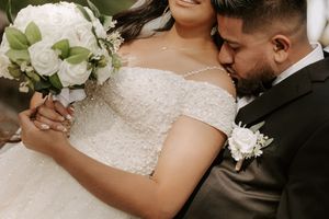 Close-up wedding photo showing a white bouquet, lace bridal gown, and boutonniere detail.