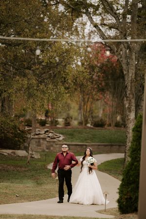 A couple walks down a winding autumn path surrounded by trees with red foliage and stone walls.