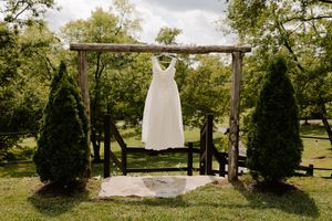 A white wedding dress hanging on a rustic wooden arch surrounded by evergreen trees in a lush green outdoor setting.
