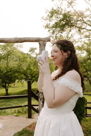 Bride in white off-shoulder dress holds a white dove outdoors at a rustic wooden fence surrounded by green trees.