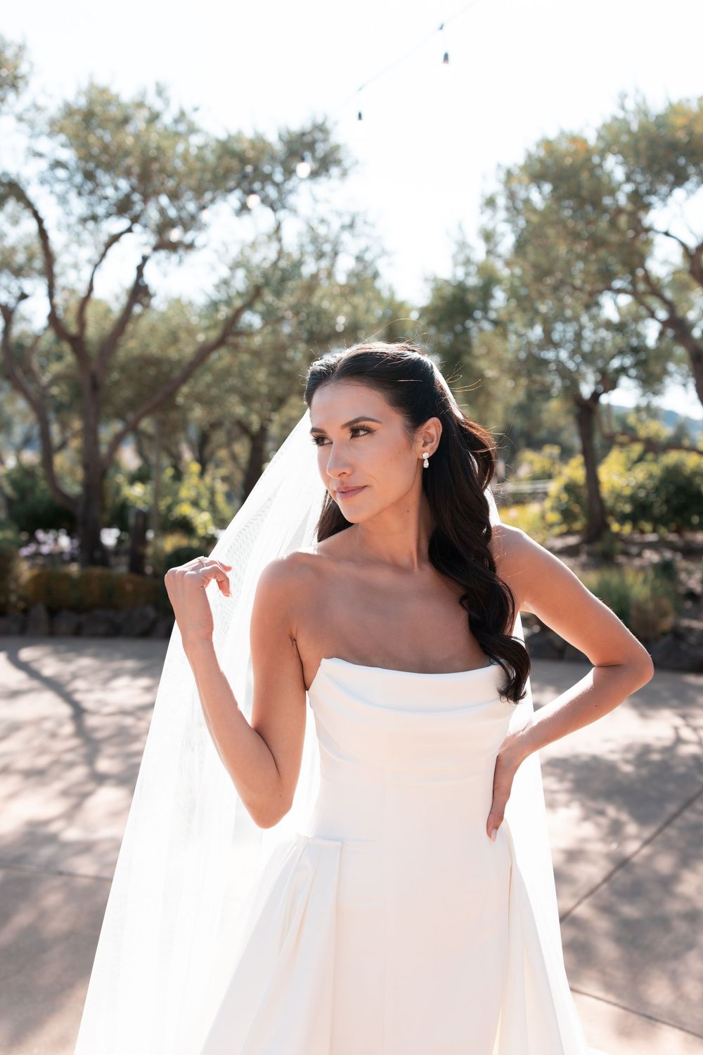 A figure in a flowing white dress poses gracefully against a backdrop of trees in an outdoor setting.