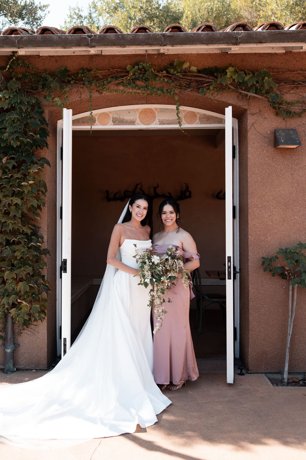 Wedding party members in pink and white dresses pose in an adobe-style doorway at a southwestern venue.