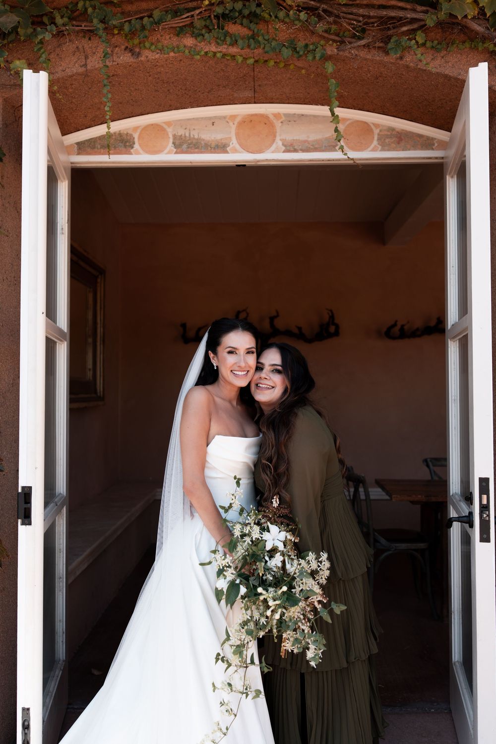 Two women stand in a doorway, one wearing a white wedding dress with cascading floral bouquet.