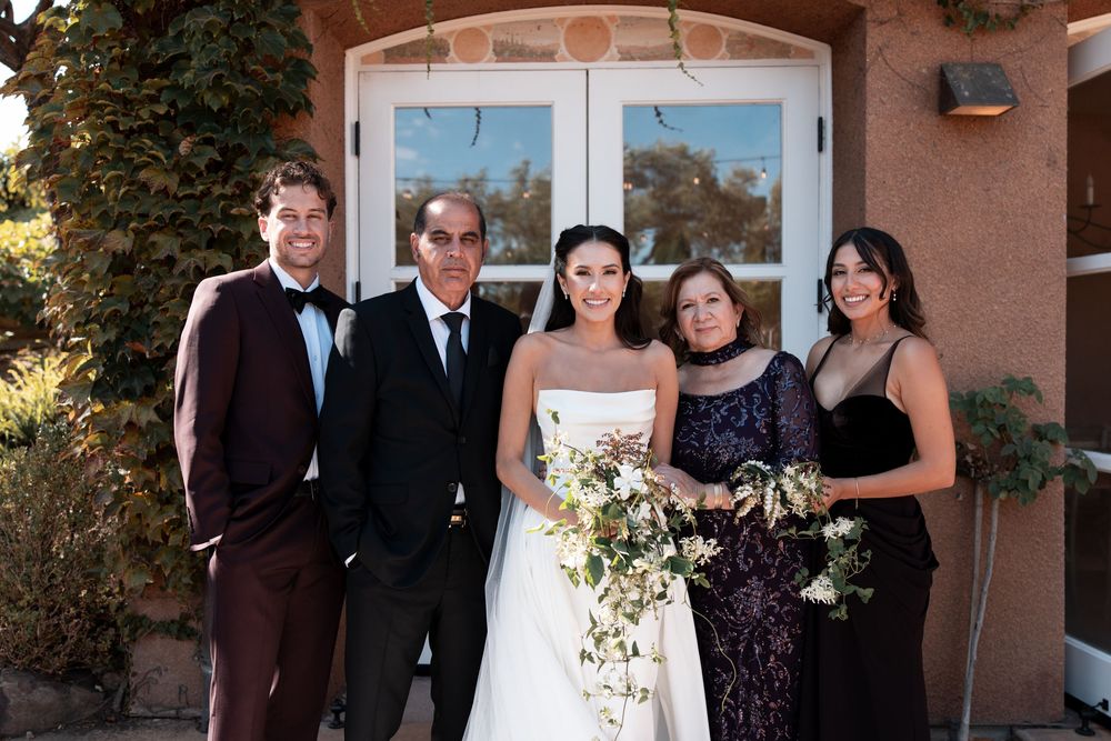 Wedding party members gather for formal photos outside a rustic adobe-style building.