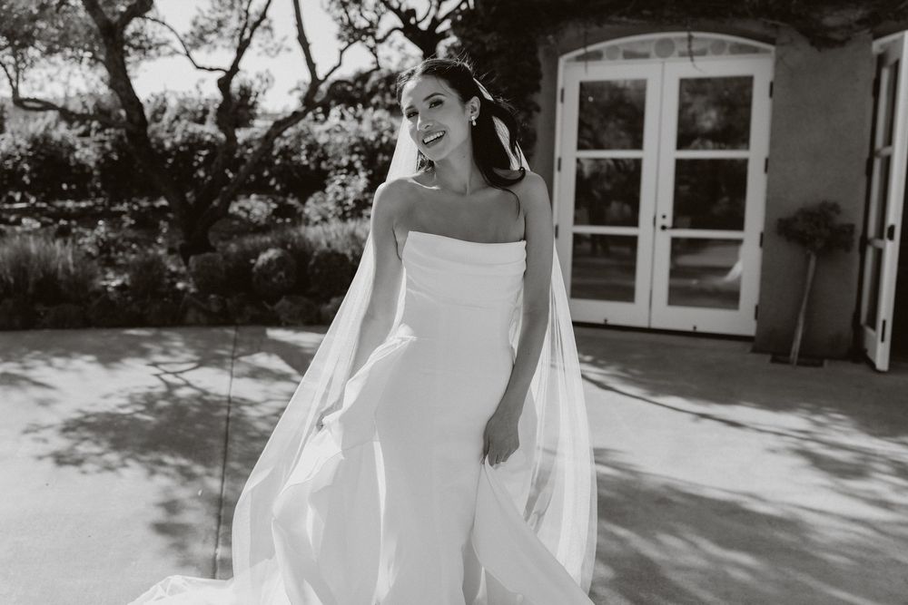 A bride in a flowing white wedding dress and veil walks gracefully through a sunlit outdoor venue.