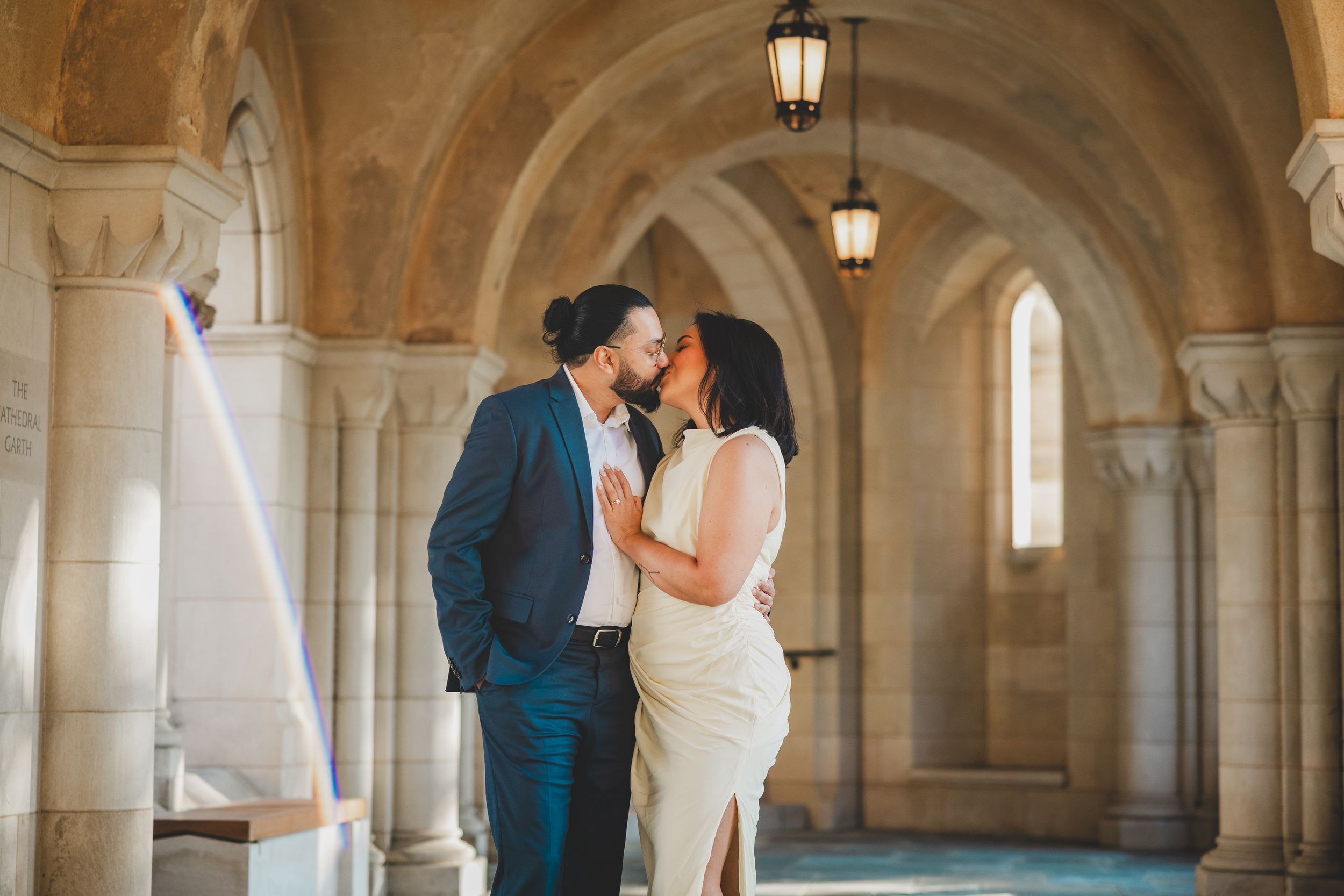 A couple shares a romantic kiss under ornate stone arches in a historic building interior.