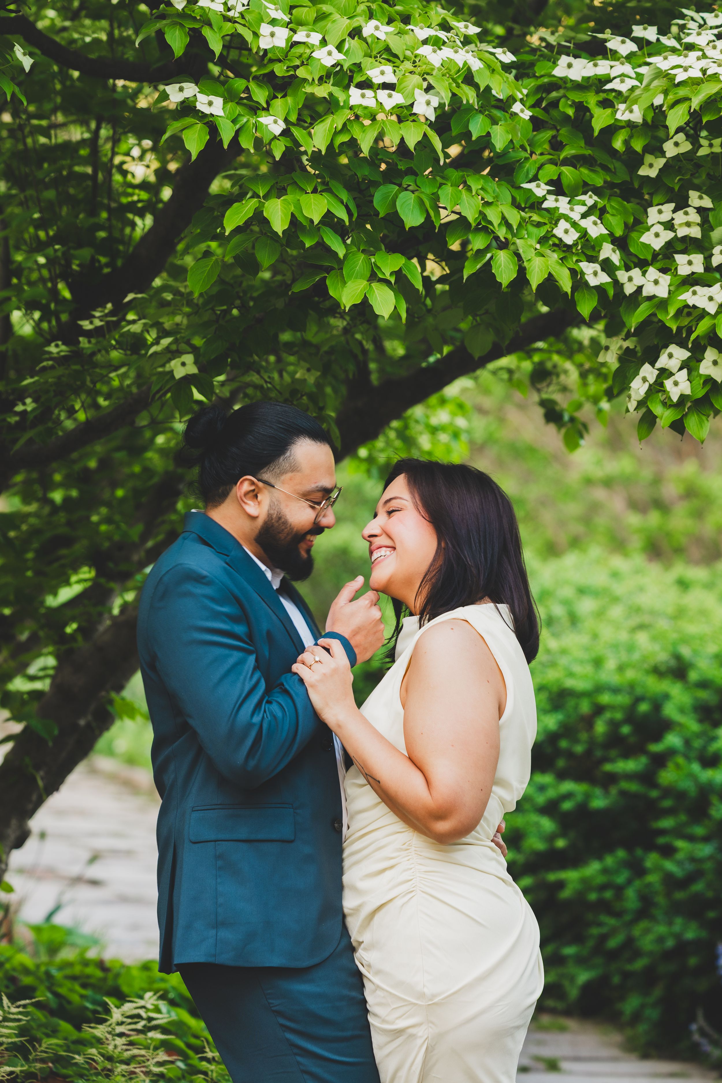 A joyful couple shares a candid laughing moment together under green tree branches.