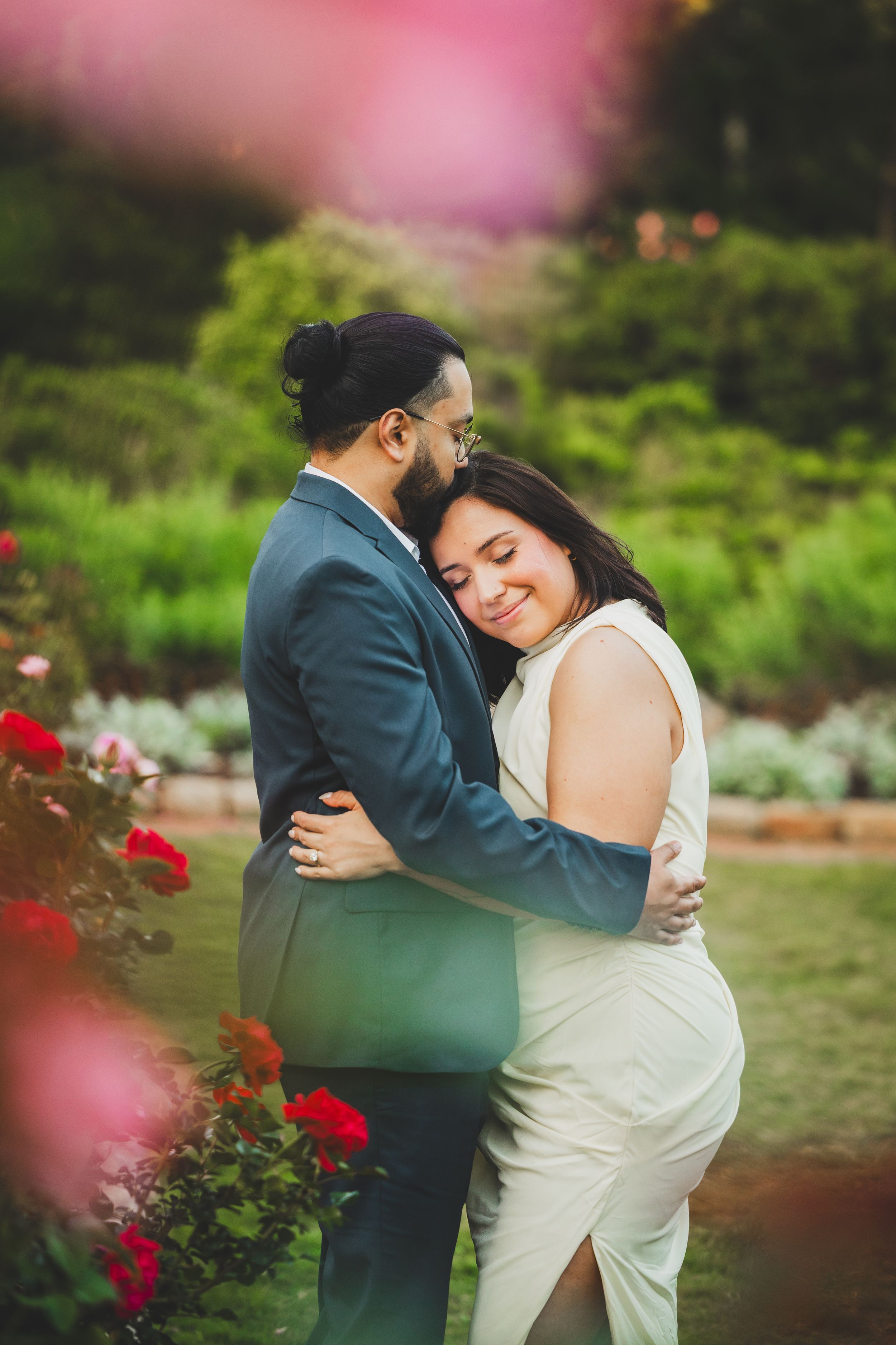 A romantic couple embraces in a garden with red roses and soft pink bokeh effects in the background.