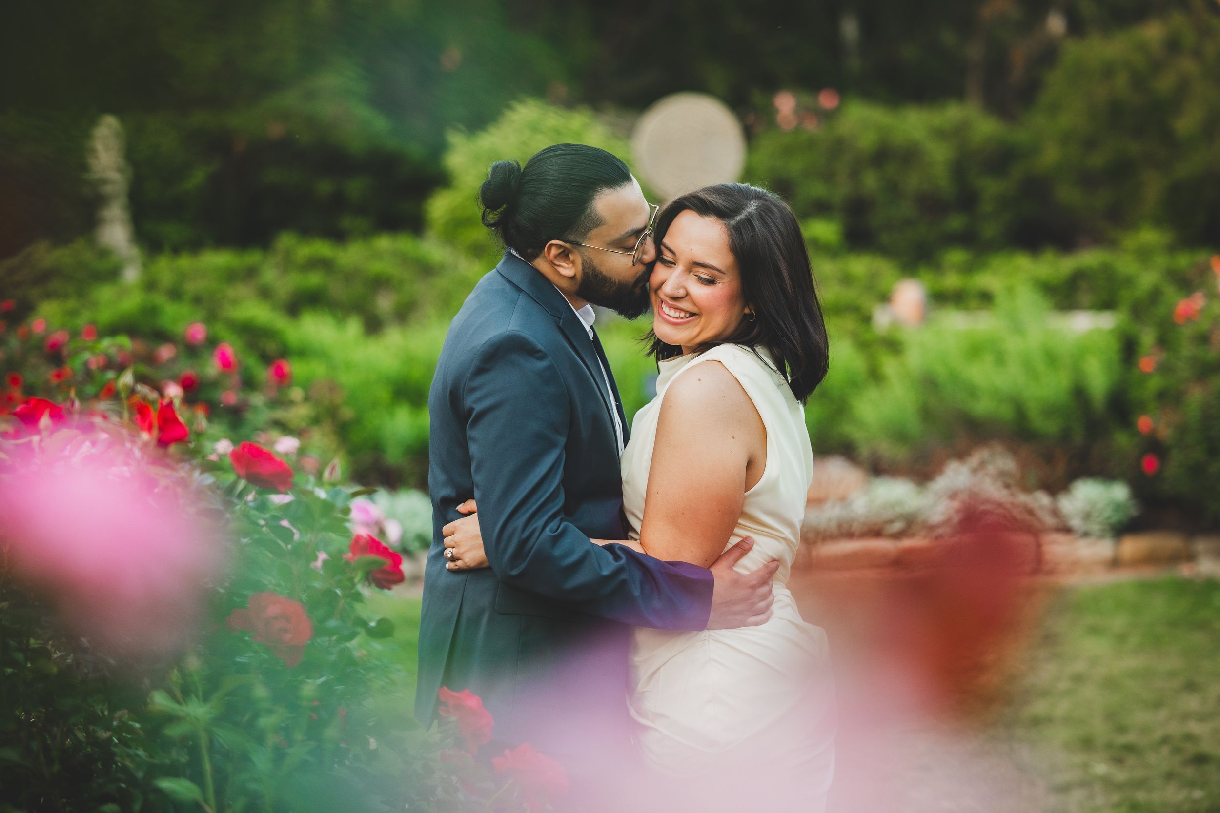 A romantic couple shares intimate moments in a lush garden setting surrounded by vibrant red roses and greenery.