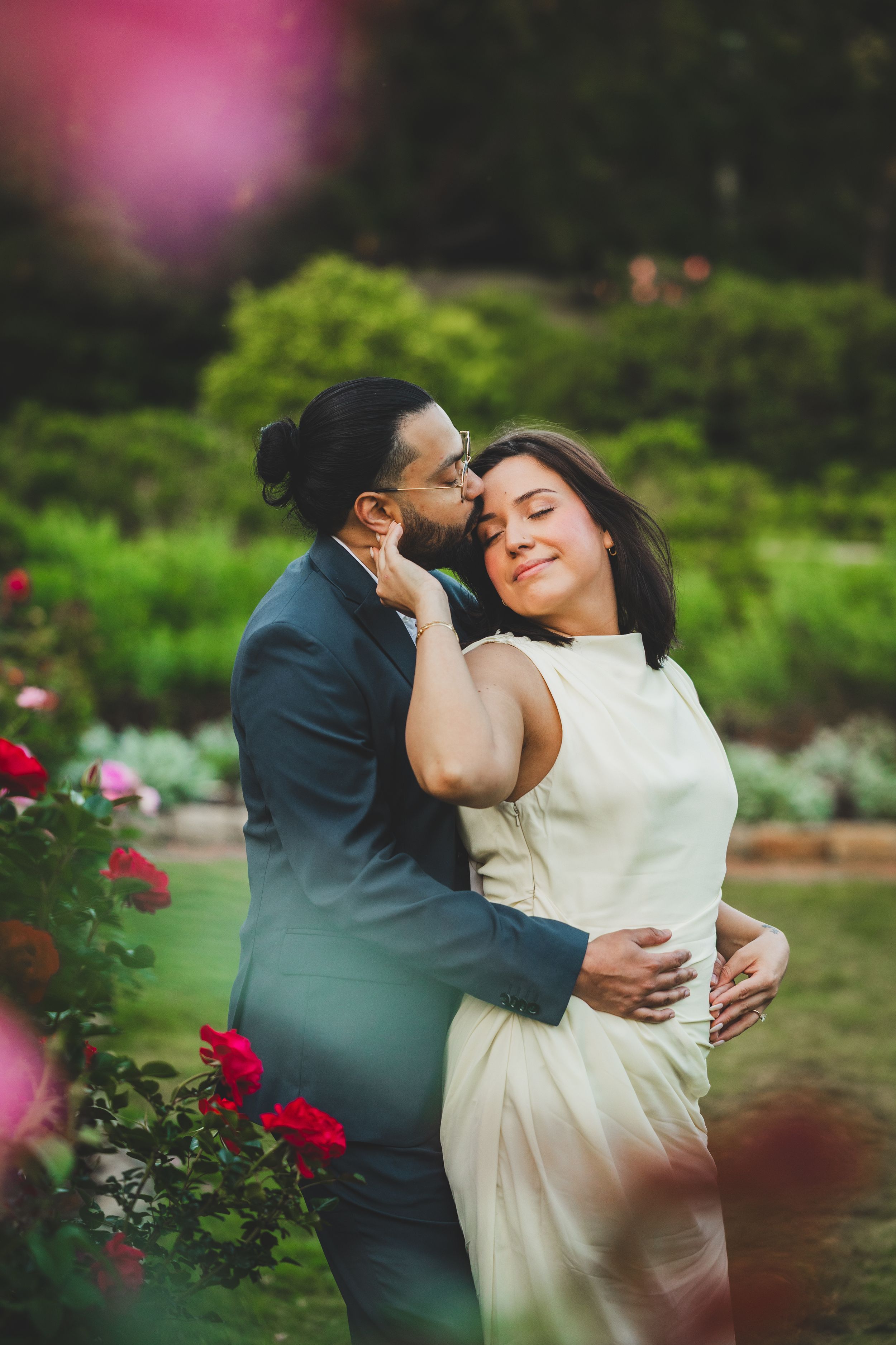 A romantic wedding couple embraces in a lush garden with pink and red roses blooming in the background.