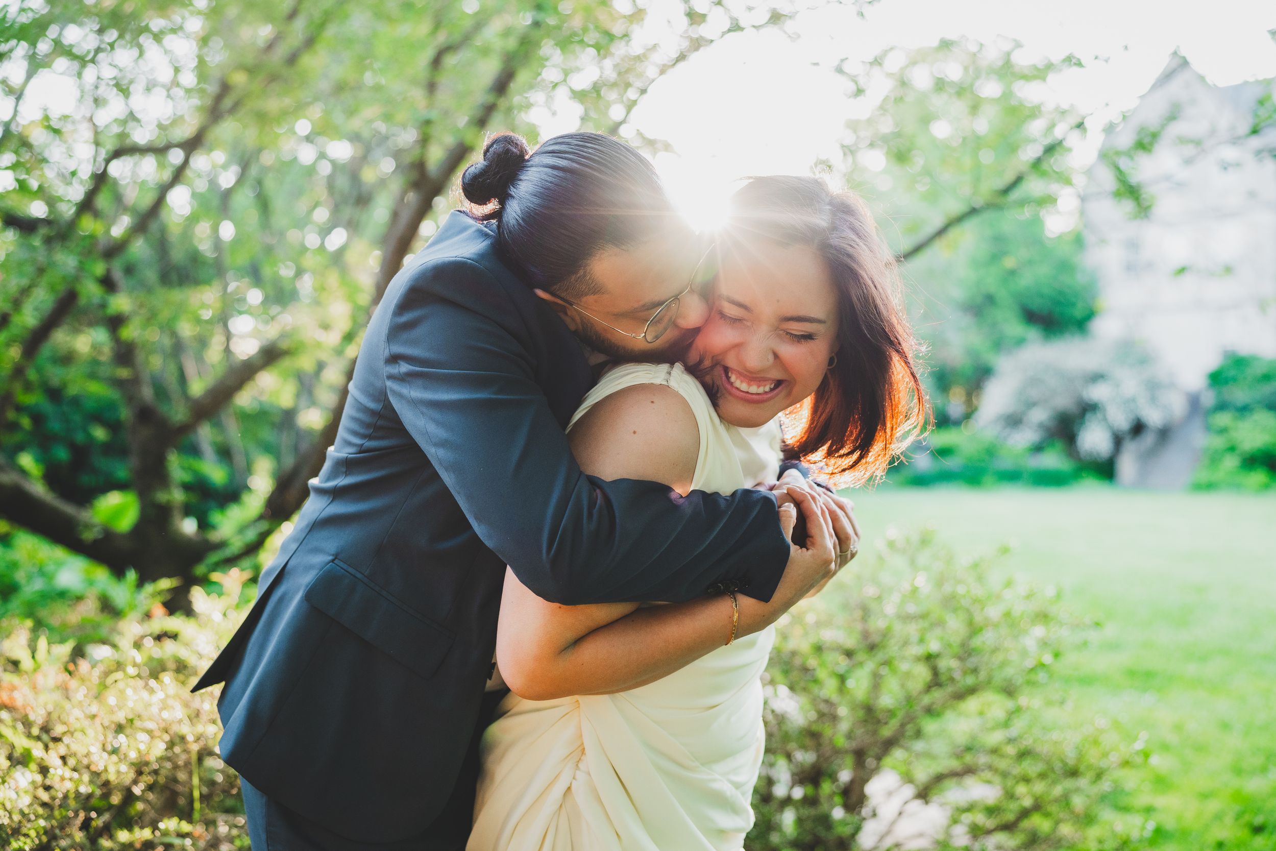 Couple embraces in a sunny garden setting with cherry blossoms blooming in the background.