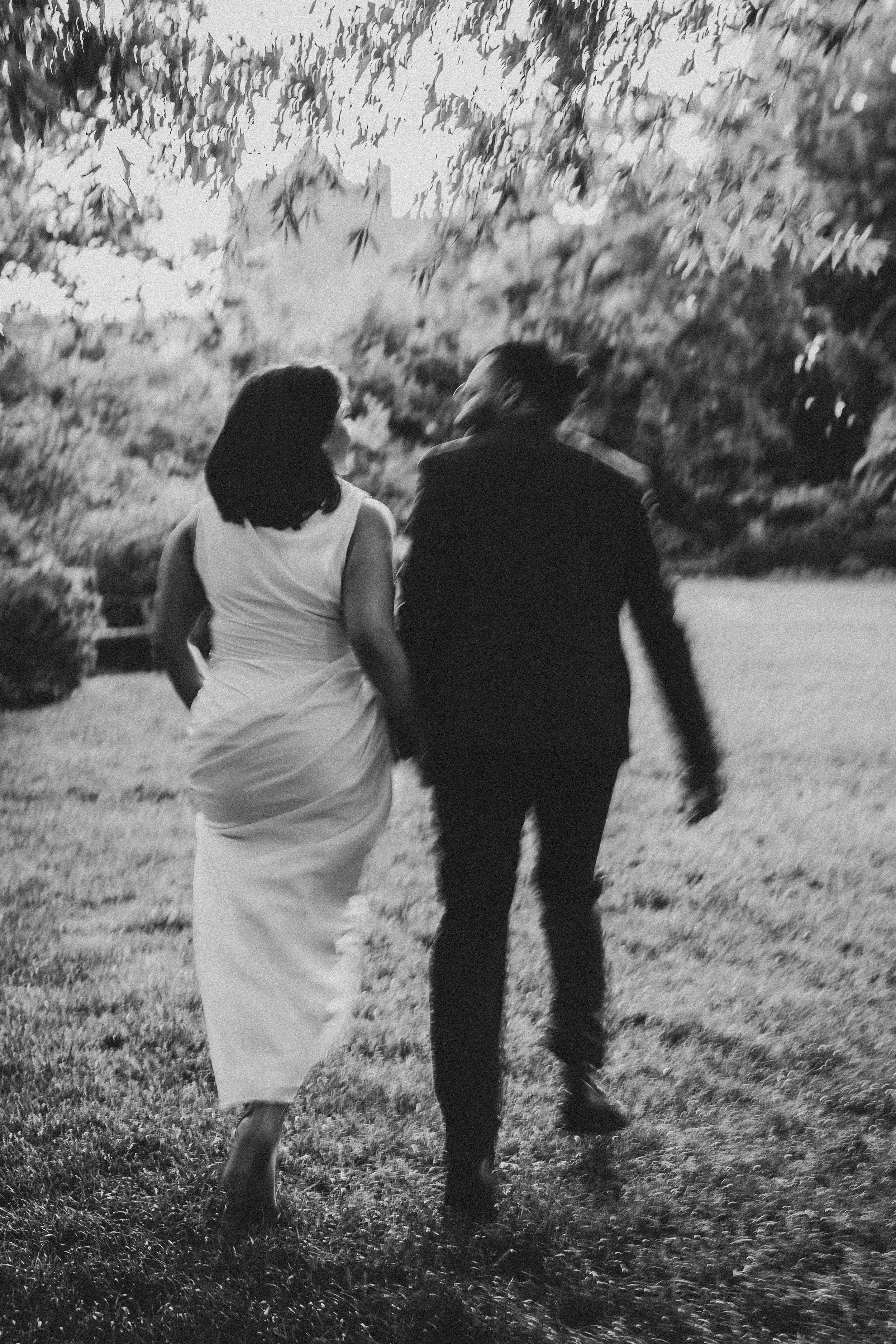 A couple walks away from camera through a garden path in black and white, with the woman in white dress and man in dark suit.